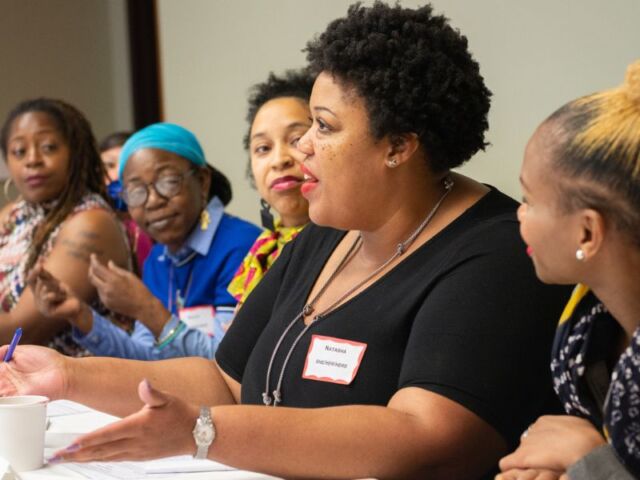 Five women are seated side-by-side along a table with papers, pens, and coffee cups scattered. Centered is a Black woman who is speaking, while the other women are attentively listening.